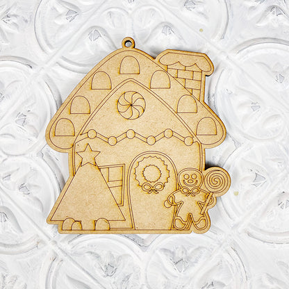 Introducing the Color Your Own Ornament from Janet's Craft Corner, a delightful wooden ornament shaped like a gingerbread house with intricate etched designs. This DIY Decoration Kit includes charming features such as a tree, wreath, candy, and a gingerbread figure holding a lollipop on a white textured surface. Perfect for adding personal touches to your home décor craft.