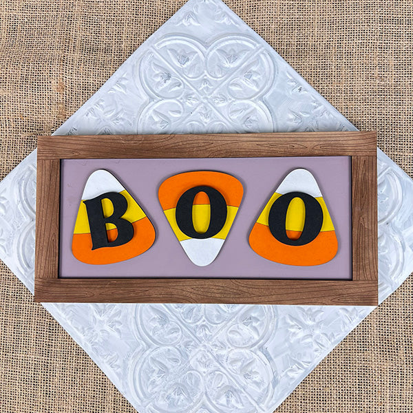 Add a festive touch to your home décor with the Boo Candy Corn Shelf Leaner from Janet's Craft Corner. This delightful piece showcases "BOO" in bold black letters on candy corn shapes, set against a light purple background and elegantly framed in wood. It rests beautifully on a white textured tile and burlap surface.