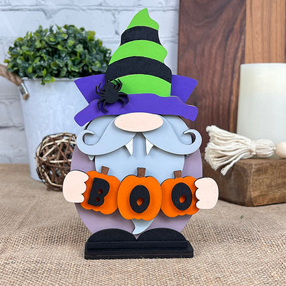 A Vampire Gnome by Janet's Craft Corner, featuring a purple and green striped hat, a white beard, and a spider holding orange letters spelling "BOO," sits on burlap beside a potted plant and decorative tassels, adding the ideal touch of spooky charm against the stark white brick wall.