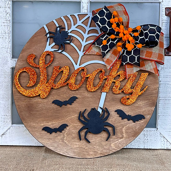 The Spooky Halloween Door Hanger from Janet's Craft Corner features a wooden circle design, embellished with the word "Spooky" in glittery orange letters, accompanied by black spiders, bats, and a web. Enhancing its charm is a plaid and spotted ribbon accented with orange berries in the top right corner. It's an ideal addition to home decor against a rustic white backdrop.