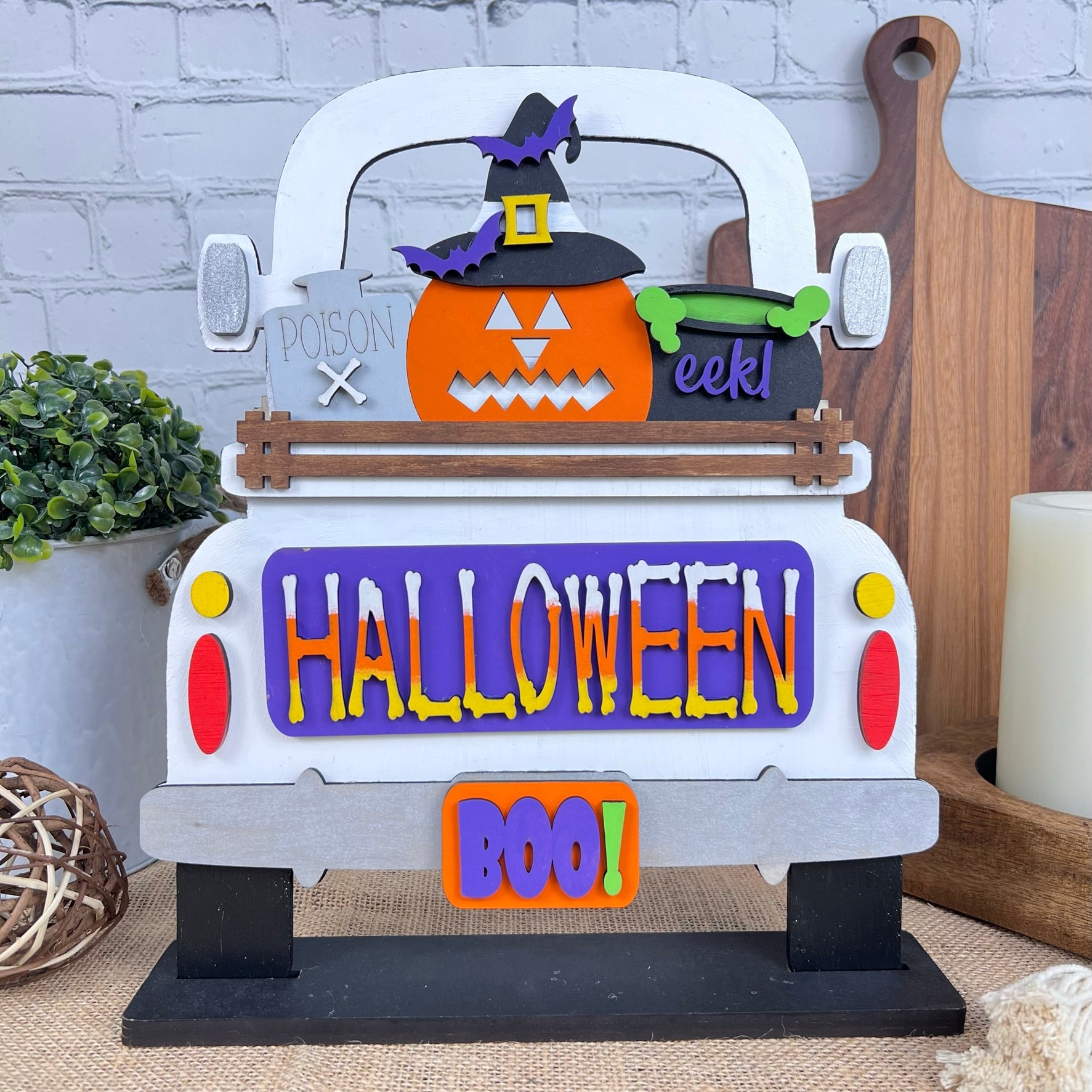 This delightful DIY home decor kit features Janet's Craft Corner's Halloween Pumpkin Interchangeable Insert, showcasing a truck with a cheerful pumpkin wearing a witch hat. The vibrant "HALLOWEEN" and "BOO!" text enhance its festive appeal. Accompanied by a plant, candle, and cutting boards set against a brick wall backdrop, it's ideal for those who enjoy interchangeable craft inserts.