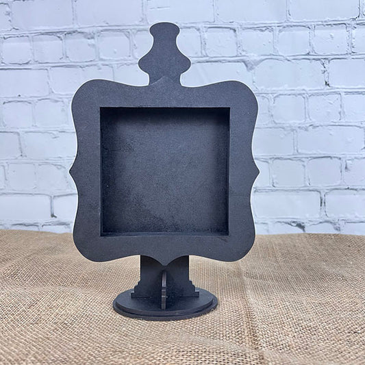 The Drop In Swappable Squares Holder DIY Craft Kit by Janet's Craft Corner is a decorative black frame with ornate edges and a stand, ideal for home décor or as a sign holder, shown on burlap before a white brick wall.