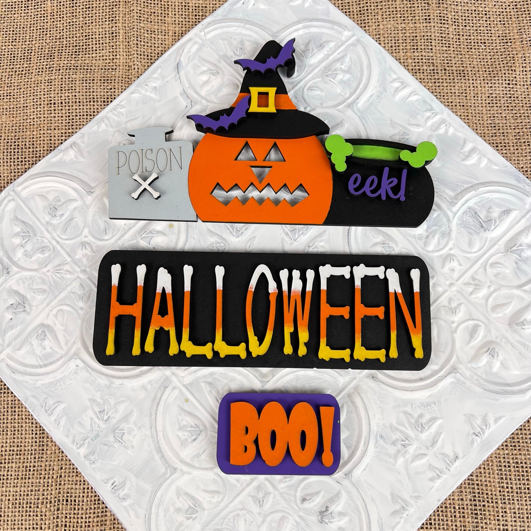 Discover the Halloween Pumpkin Interchangeable Insert from Janet's Craft Corner, a DIY home decor kit that features a festive sign with interchangeable craft inserts. It includes a pumpkin sporting a witch hat, an "eek!" marked cauldron, and a poison label. The textured background highlights the words "HALLOWEEN" and "BOO!", perfect for adding charm to your Halloween decor.