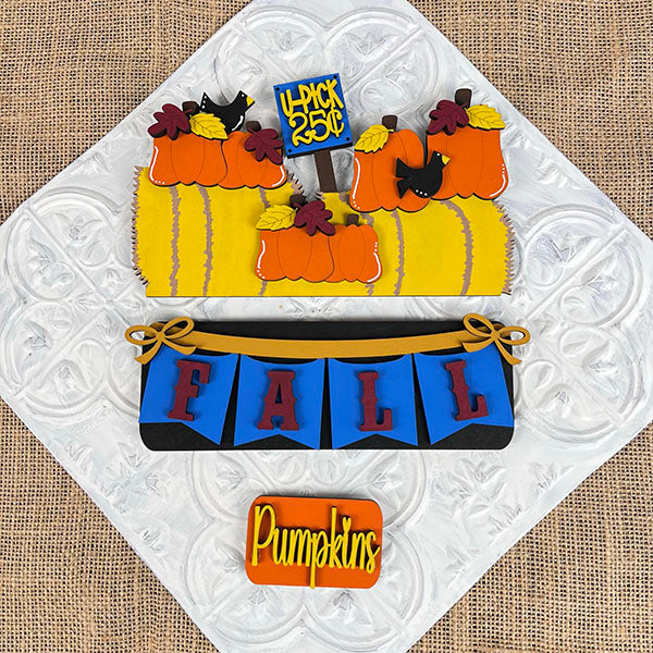 A set of beautifully decorated cookies displayed on a woven mat captures the essence of fall décor with designs featuring pumpkins, hay bales, and crows. One cookie displays the message "4-Pick 5 for 25¢" in a cozy autumn atmosphere, perfectly embodying the style of Janet's Craft Corner's Fall Hay Bale Interchangeable Insert - Painted. This scene brings an interchangeable home decor touch to your seasonal festivities.