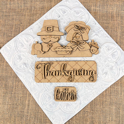 Introducing the Pilgrim Interchangeable Insert from Janet's Craft Corner: a charming decoration depicting a carved pilgrim man and woman with a pie. Beneath them, signs display "Thanksgiving" and "Gather." The textured white and burlap backdrop is ideal for use in your Thanksgiving decor or as part of your DIY home decor kit.