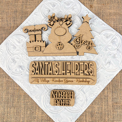 Janet's Craft Corner presents the "Santa's Helpers Interchangeable Insert" DIY home décor kit, showcasing wooden cutouts on a textured background to bring charm to your holiday scene. It features a chimney, an antlered snowman, and a tree with the word "Joy," along with signs reading "Santa's Helpers" and "North Pole." This kit is perfect for festive creativity with its interchangeable inserts.