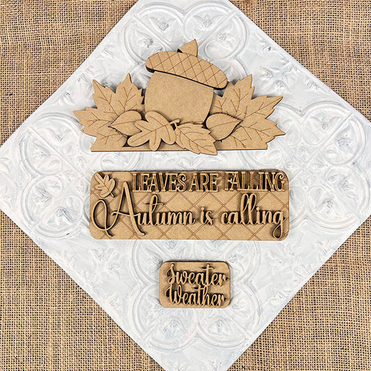 The Autumn Is Calling Interchangeable Insert, a DIY home decor kit from Janet's Craft Corner, showcases hand-painted autumn-themed designs on a textured white and burlap background. The central pieces highlight an acorn surrounded by leaves with the phrase "Leaves are falling, Autumn is calling," accompanied by a smaller piece featuring "Sweater weather." Ideal for interchangeable insert collections or crafting projects.