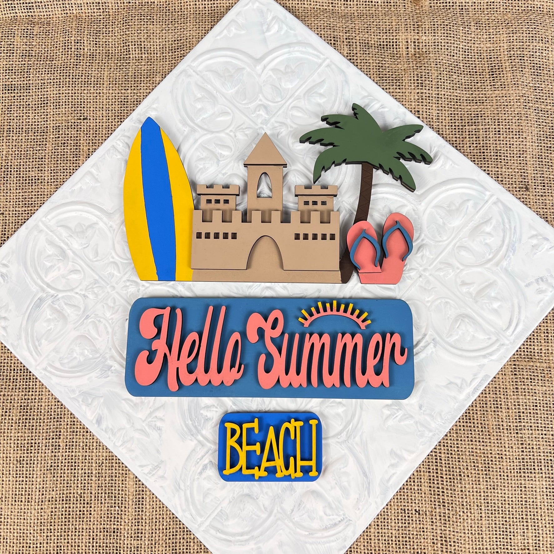 The Summer Beach Interchangeable Insert from Janet's Craft Corner features a textured decorative sign with a sandcastle, surfboard, palm tree, and flip-flops. It displays "Hello Summer" alongside a sun graphic above the bold word "BEACH," making it an ideal DIY home decor kit to add style to your space!