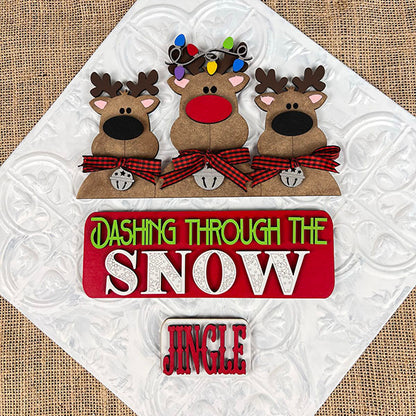 Janet's Craft Corner presents the Reindeer Interchangeable Insert DIY home decor kit, featuring three charming cartoon reindeer with red noses, plaid bow ties, antlers, and bells. This delightful piece adds a festive touch to your holiday sign with the message "Dashing Through The Snow" and "Jingle" at the bottom, making it ideal for creating a warm and inviting seasonal ambiance.