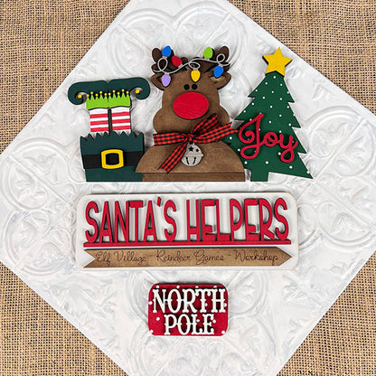 On a white surface, a festive display features a light-adorned reindeer, an elf shoe, and a green Christmas tree with the word "Joy." A sign labeled "Santa's Helpers" from Janet's Craft Corner includes an interchangeable insert for your home decor. Adding to the scene are signs that read "Elf Village Reindeer Games Workshop" and "North Pole," both crafted on burlap.