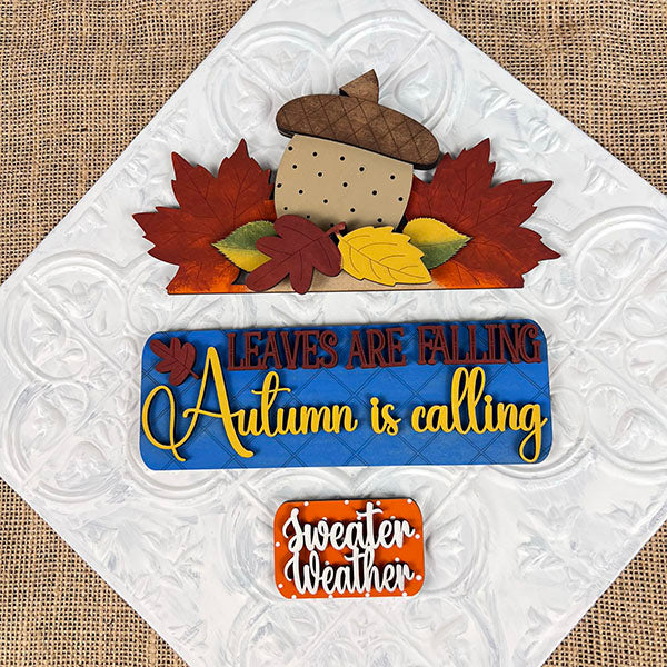 Autumn-themed decorative signs from Janet's Craft Corner, featuring the DIY home decor kit "Autumn Is Calling Interchangeable Insert." The top sign showcases an acorn with leaves and the phrase "Leaves are falling," while the customizable middle sign states, "Autumn is calling," providing an interchangeable touch. The bottom sign reads, "Sweater weather," embellished with leaves.