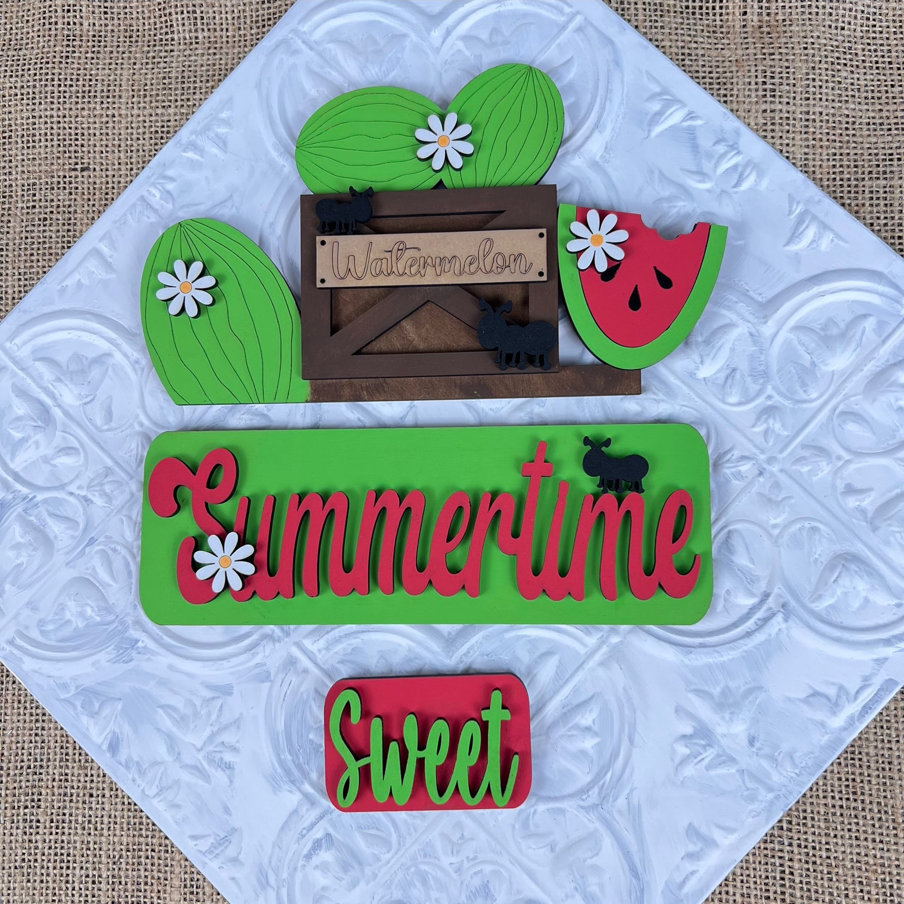 Decorative pieces from Janet's Craft Corner are arranged on a textured white square with a burlap background. The Summertime Watermelon Interchangeable Insert features a wooden watermelon sign alongside green and red "Summertime" and "Sweet" cutouts, small daisies, and hand-painted black elephant shapes for the perfect summer flair.