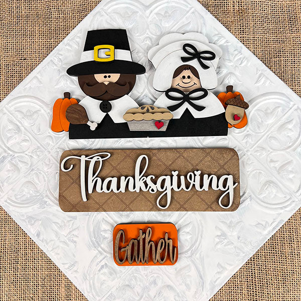 The Pilgrim Interchangeable Insert from Janet's Craft Corner is a DIY home decor kit that features a decorative design with a pilgrim couple, pumpkins, a pie, and an acorn. The word "Thanksgiving" is prominently displayed with "Gather" beneath it on a burlap-textured background, ideal for refreshing your Thanksgiving decor or creating unique craft projects.