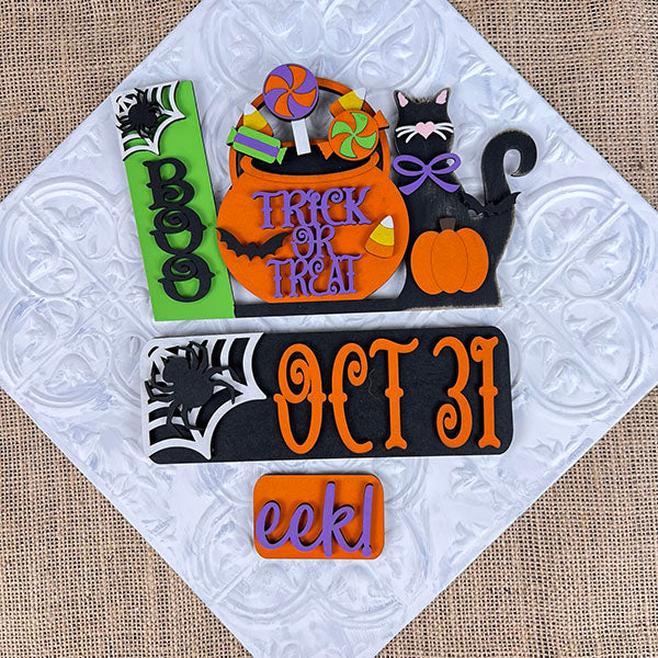 Janet's Craft Corner's Oct 31 Interchangeable Insert DIY home decor kit features Halloween-themed designs, including a hand-painted tombstone with "BOO" and a pumpkin with "TRICK OR TREAT," all artfully arranged on a textured white surface, creating a delightful display reminiscent of interchangeable home decor sets.