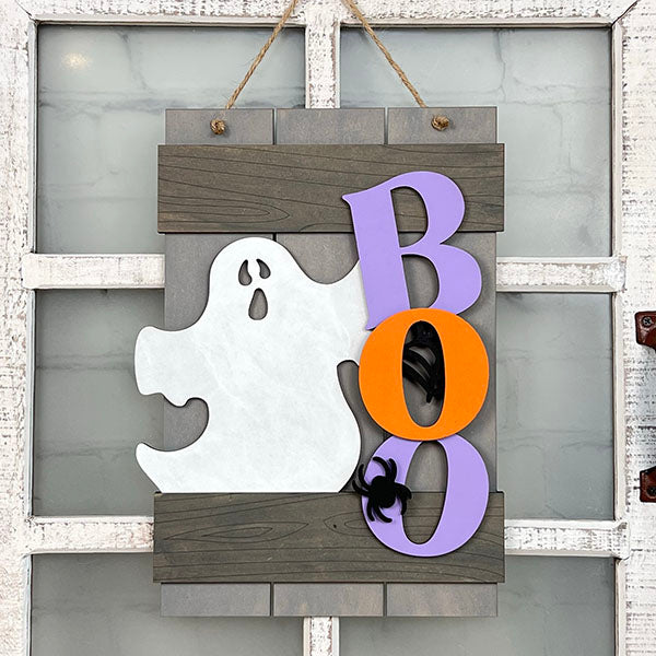 The Boo Fence Door Hanger from Janet's Craft Corner showcases a white ghost silhouette complemented by the word "BOO" crafted in vibrant purple and orange letters set against a wooden backdrop, with black spiders enhancing the spooky vibe, making it an ideal piece for Halloween home décor.