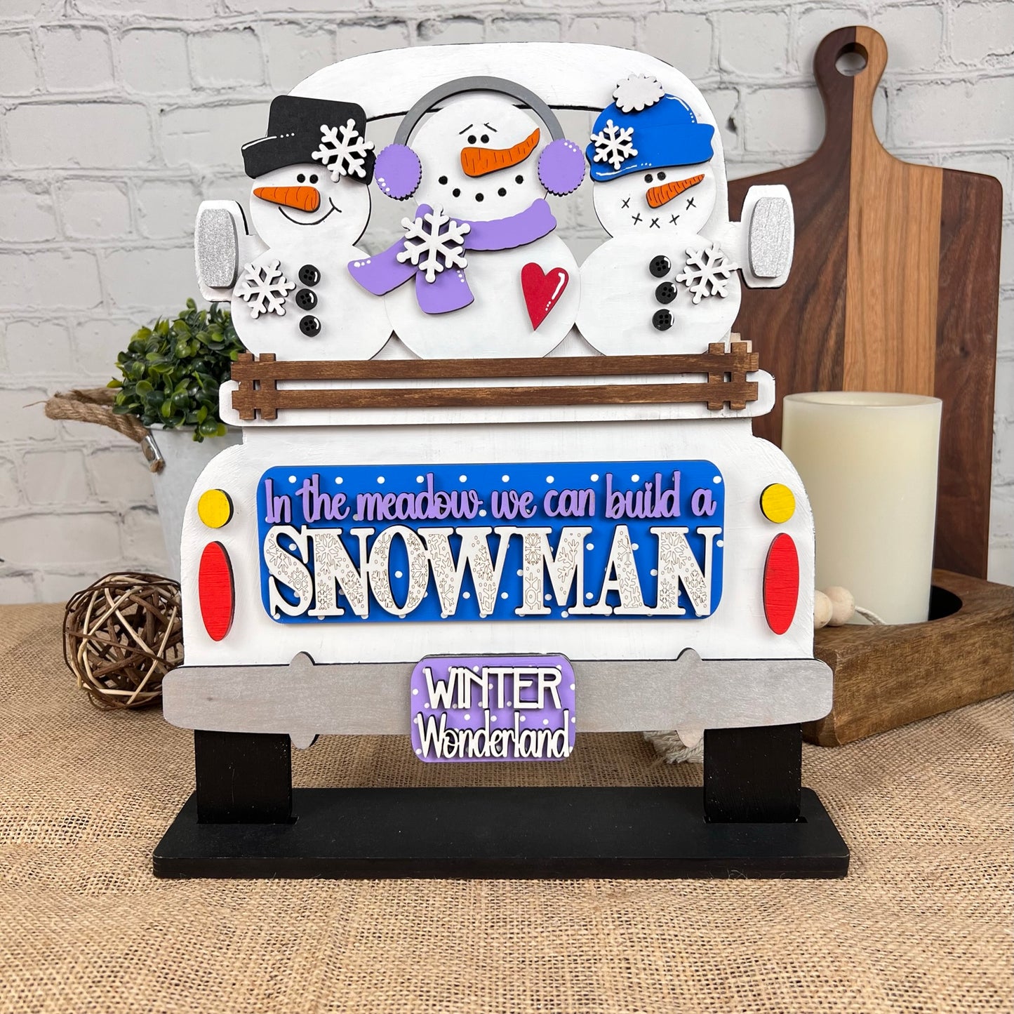 Introducing the Antique Truck Base with Interchangeable Insert by Janet's Craft Corner—a delightful DIY home decor kit. This decorative wooden sign features an antique truck design adorned with charming snowman elements and includes inscriptions like "In the meadow we can build a SNOWMAN" and "WINTER Wonderland." The backdrop, showcasing a candle and chopping board on a burlap surface, adds a cozy ambiance to your space.