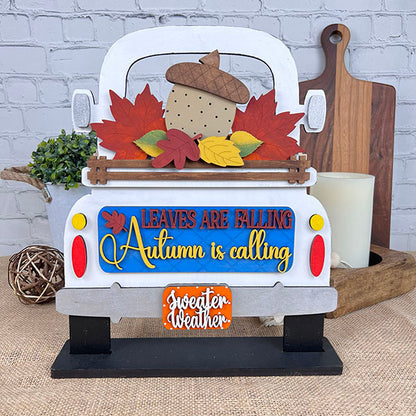 Janet's Craft Corner offers the "Autumn Is Calling Interchangeable Insert," a DIY home decor kit, featuring a decorative piece styled like the back of a truck with vibrant leaves and an acorn. The sign displays the message "LEAVES ARE FALLING, Autumn is Calling, sweater weather" and includes an interchangeable insert. It is beautifully set against a brick wall with wooden boards and a candle in the background.