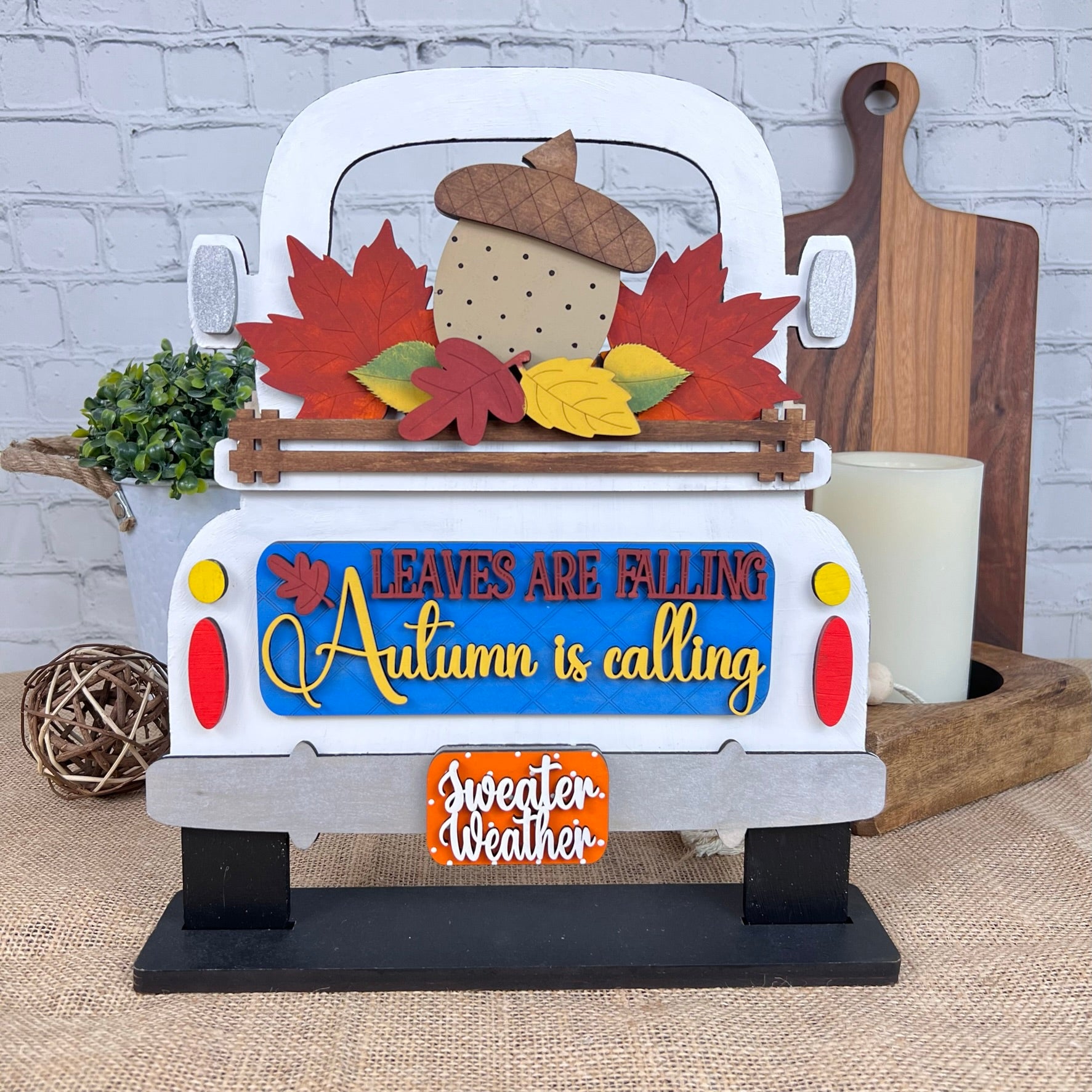 A rustic DIY home decor kit from Janet's Craft Corner features an antique truck base with interchangeable inserts, creatively painted in an autumn theme. The text on the insert says, "Leaves are falling, Autumn is calling, Sweeter weather," and it's charmingly adorned with leaves and an acorn for a seasonal touch.