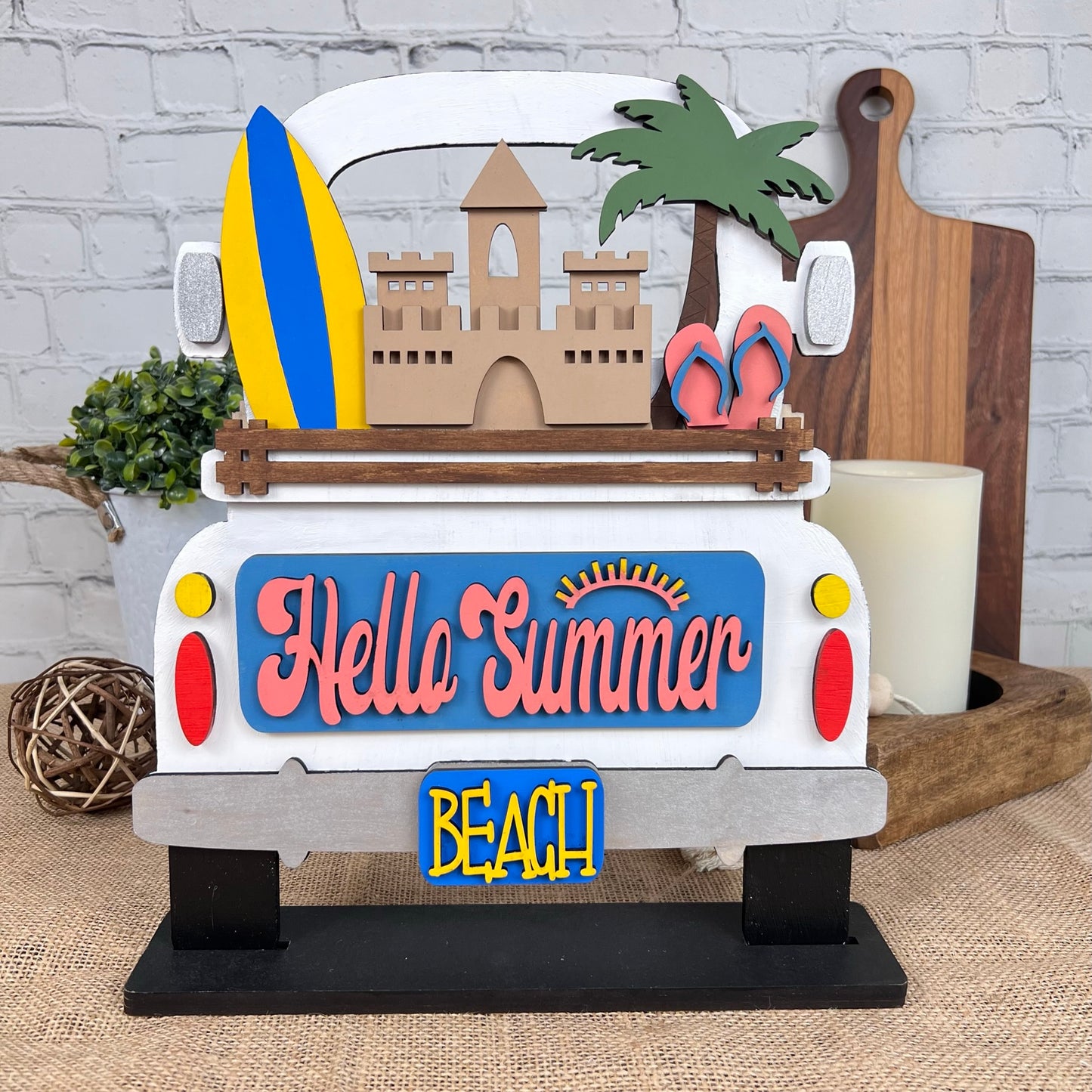 The Summer Beach Interchangeable Insert by Janet's Craft Corner is a hand-painted DIY home decor kit featuring a decorative sign that resembles the rear of a truck with a surfboard, sandcastle, palm tree, and flip-flops. The vibrant "Hello Summer" text shines above the "BEACH" license plate, making it perfect for infusing your home with summer vibes among plants and candles.