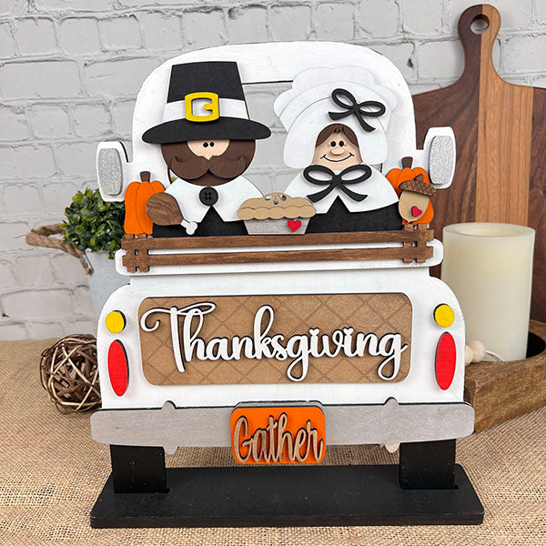 The Pilgrim Interchangeable Insert from Janet's Craft Corner, a DIY home decor kit, features a charming pilgrim couple in a vintage truck filled with pumpkins and a pie. Displaying the words "Thanksgiving" and "Gather," this piece is set against a white brick backdrop with autumn accents, making it the ideal addition to enhance any Thanksgiving setting.