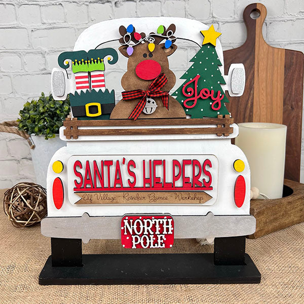 Add a touch of charm to your wooden shelf with this festive decoration from Janet's Craft Corner. The Santa's Helpers Interchangeable Insert features a vintage truck carrying a reindeer, wrapped presents, and a Christmas tree. The "North Pole" license plate adds the final touch to this perfect hand-painted ornament.