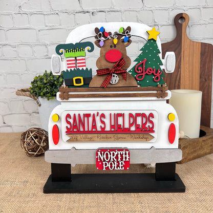 A festive decoration from Janet's Craft Corner features an antique truck base with a reindeer adorned with ornaments, a Christmas tree, and gift boxes. The truck bears the phrases "SANTA'S HELPERS" and "NORTH POLE" and rests on a burlap-covered table.