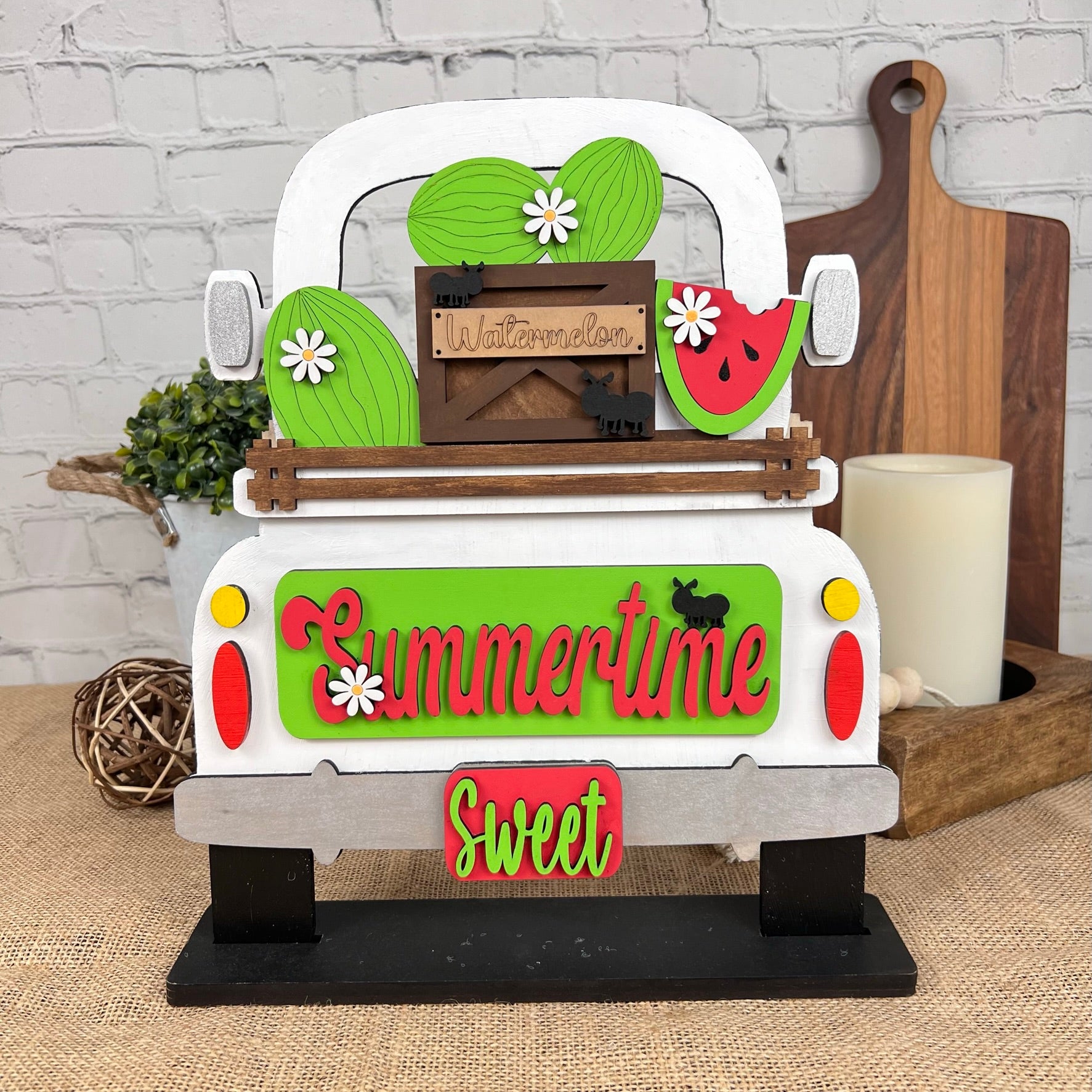 Introduce a touch of summer charm with Janet's Craft Corner's "Antique Truck Base with Interchangeable Insert" DIY home decor kit. This decorative wooden piece features a vintage truck carrying a crate of watermelons, adorned with daisies and vibrant colors. The tailgate is elegantly inscribed with "Summertime Sweet," while rustic kitchen accents complement the scene against a white brick wall backdrop—ideal for your next hand-painted craft project.