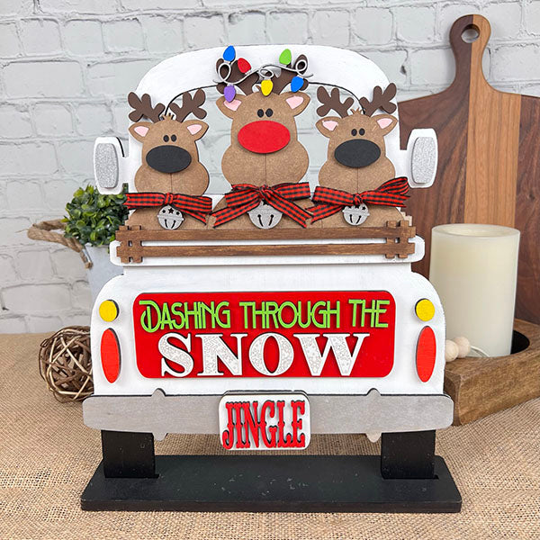 Introducing the charming Reindeer Interchangeable Insert from Janet's Craft Corner. This DIY home decor kit features three cartoon reindeer adorned with bows, nestled in a vintage truck, with a sign that reads "Dashing Through the Snow" and "Jingle" below. Perfectly set against a backdrop of candles and a rustic wall, it's an ideal way to add charm to your festive celebrations!