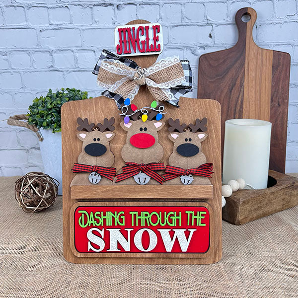 This delightful DIY home decor kit from Janet's Craft Corner, known as the Reindeer Interchangeable Insert, showcases three reindeer with red noses and vibrant lights. A "Jingle" sign adorns the top, while "Dashing Through the Snow" accents the bottom. The scene is set against a white brick wall, with a candle and wooden boards completing the background ambiance.