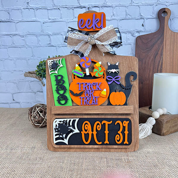 This captivating DIY craft kit, the Oct 31 Interchangeable Insert from Janet's Craft Corner, is a hand-painted wooden Halloween sign. It features "Boo," "Trick or Treat," candy, bat, black cat, pumpkin, and "Oct 31" in vibrant orange and black colors. Topped with a bow, it makes for perfect interchangeable home decor against a brick wall backdrop.