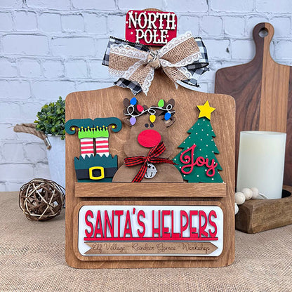 Introducing the "Santa's Helpers Interchangeable Insert - Painted" from Janet's Craft Corner, a delightful wooden sign adorned with hand-painted North Pole-themed ornaments. It features charming elements such as a reindeer, elf shoes, and a Christmas tree, topped off with "Santa's Helpers" text. An eye-catching bow and checkered fabric accent against a brick wall background make this piece perfect for festive home decor.