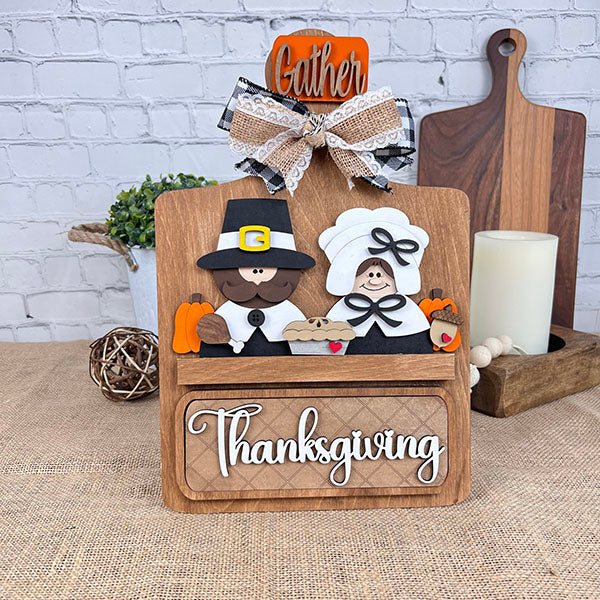 This DIY home decor kit from Janet's Craft Corner, called the Pilgrim Interchangeable Insert, features a charming pilgrim couple with pumpkins, pies, and a turkey. Topped with a "Gather" sign and lace bow, it has "Thanksgiving" written below. Perfect for crafters, this versatile kit offers interchangeable inserts for use throughout the year.