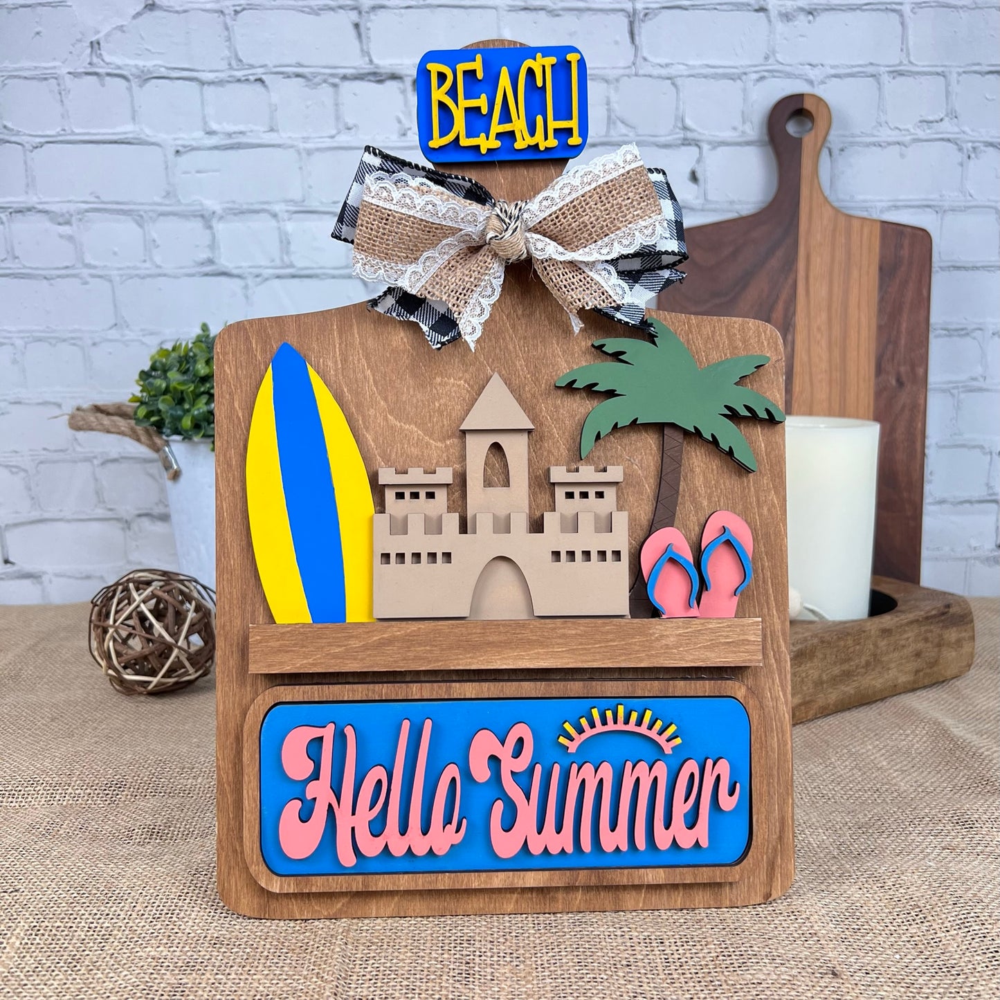 A wooden decorative sign by Janet's Craft Corner, from their Summer Beach Interchangeable Insert DIY home decor kit, is set against a brick wall. It features delightful hand-painted images of a sandcastle, surfboard, palm tree, and flip-flops. The scene exudes interchangeable home decor charm with signs reading "Hello Summer" and "Beach." To complete the setup, cutting boards and a candle are also included.