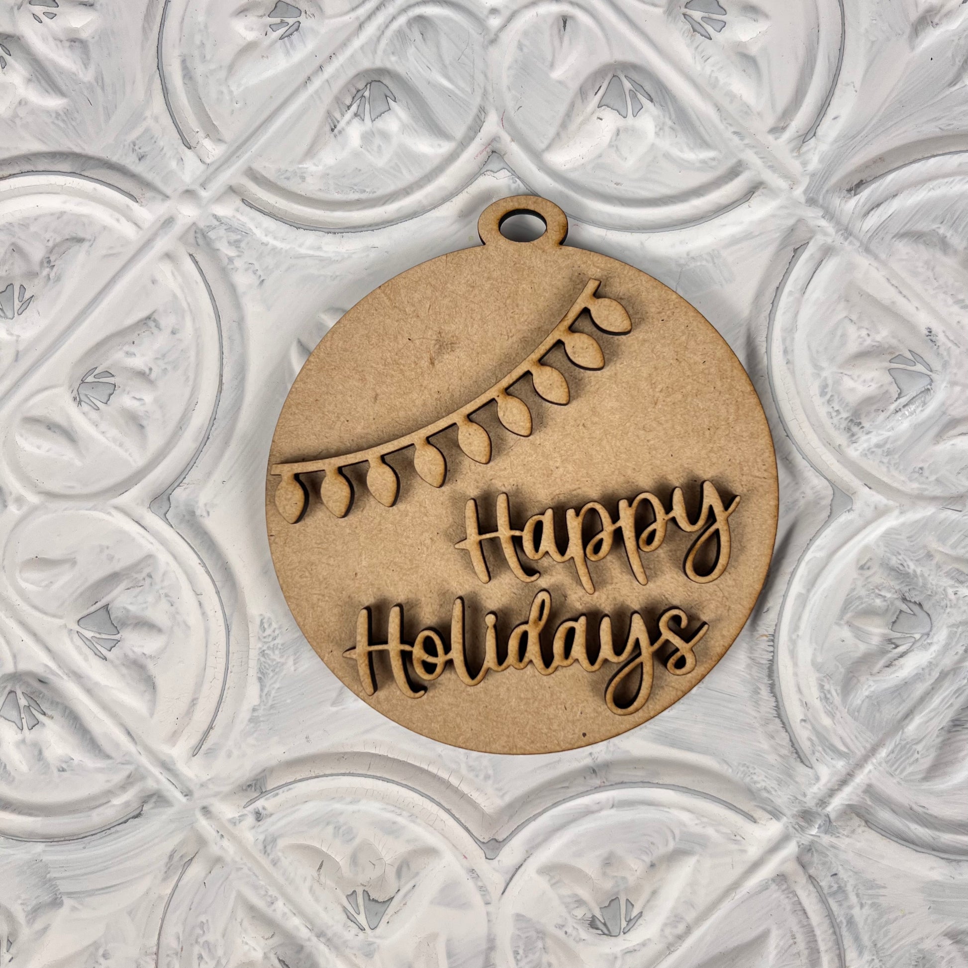 A Round Christmas Ornament from Janet's Craft Corner rests on a textured white surface. This delightful piece, an ideal choice for a DIY decoration kit, showcases cut-out text that says "Happy Holidays" with an elegant string of lights arching above the joyful message.