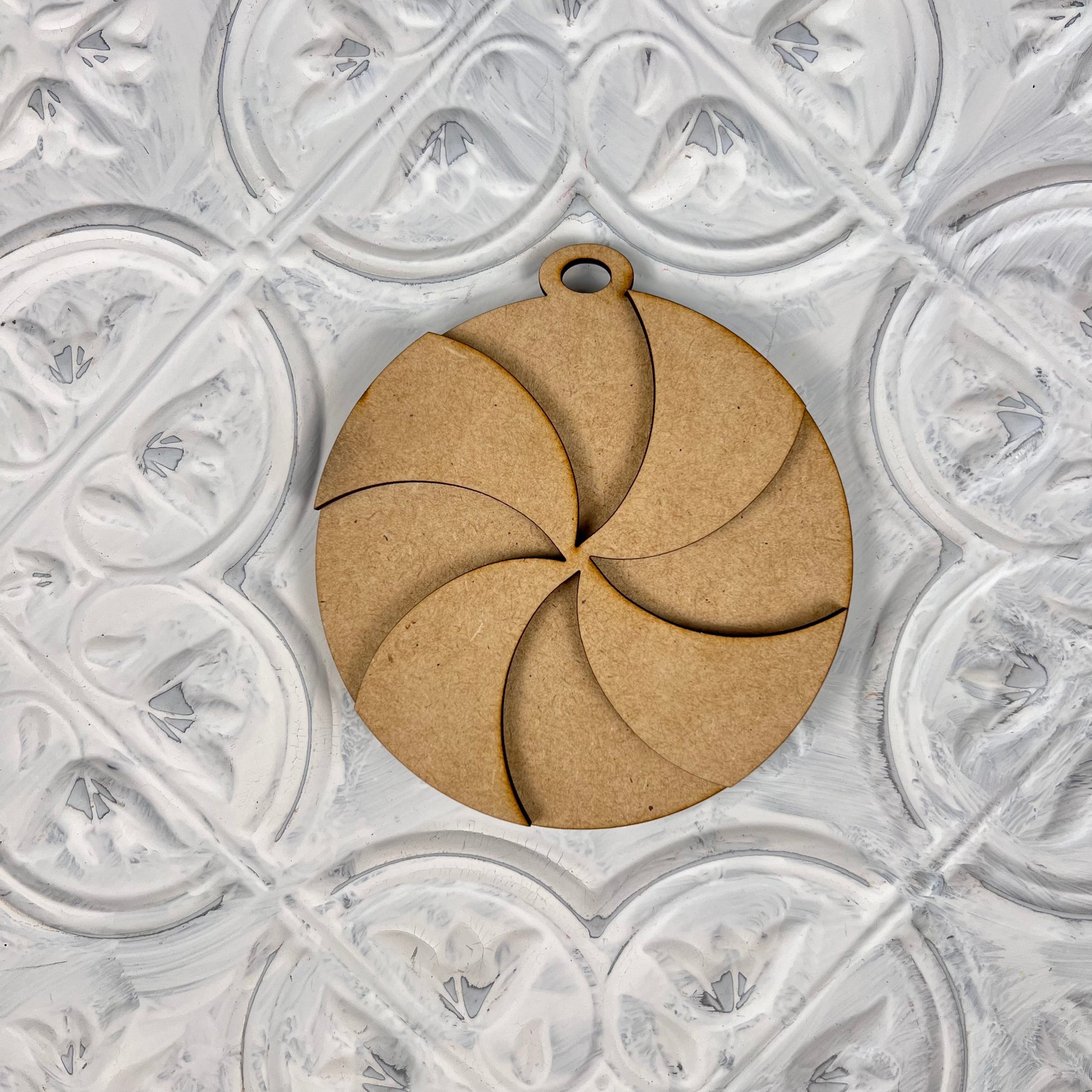 A round ornament from Janet's Craft Corner, featuring a swirl pattern, rests on an embossed white surface with a floral design. Ideal for your festive decorations, this Christmas ornament includes a small loop at the top for easy hanging.