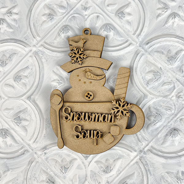 The Snowman Soup Ornament from Janet's Craft Corner is a charming holiday décor piece. This DIY craft kit includes a wooden snowman with a top hat, expressive eyes, button mouth, snowflakes, the text "Snowman Soup," and comes with a spoon and marshmallows on a textured white background.