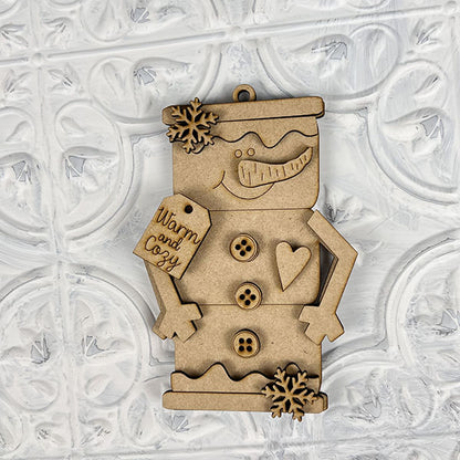 Create the perfect holiday atmosphere with a DIY decoration kit from Janet's Craft Corner, featuring wooden snowman ornaments adorned with smiling faces, buttons, heart shapes, and whimsical snowflake accents. The design is completed with a tag that reads "Warm and Cozy," set against an embossed white patterned background.