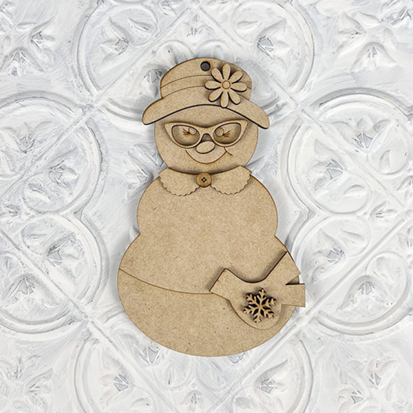 The Vintage Snow Couple Ornaments from Janet's Craft Corner are charming DIY decoration kits that feature a wooden snowman adorned with a flower-topped hat, glasses, and a scarf. One twig-like hand holds a small snowflake, adding to its vintage appeal. The textured white background evokes the look of handcrafted ornaments from days gone by.