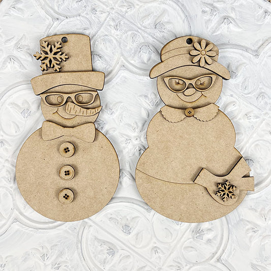 Crafted by Janet's Craft Corner, the Vintage Snow Couple Ornaments from the DIY Decoration Kits feature two intricately carved figures—a snowman and a snowwoman. They stand against a textured white backdrop, adorned with detailed buttons, glasses, and decorative hats. The snowwoman elegantly holds a small bird embellished with a snowflake, echoing the charm of lovingly painted ornaments.