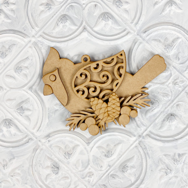 This elegant ornament from Janet's Craft Corner's DIY Decoration Kits showcases a cardinal with detailed scrollwork on its wing, poised atop pinecones and branches. Ideal for holiday décor, it stands out wonderfully against a textured white background.