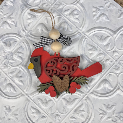 This Cardinal Ornament from Janet's Craft Corner, featured in the DIY Decoration Kits collection, is ideal for holiday décor. It showcases a yellow beak and detailed cut-out designs. Adorned with a pinecone, berries, and wooden beads topped with a ribbon, it shines elegantly against a textured white background.