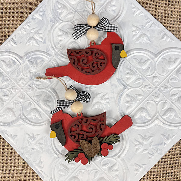 The Cardinal Ornament from Janet's Craft Corner showcases detailed red cardinal designs with intricate patterns and black-and-white checkered bows. Ideal for holiday décor, these ornaments are enhanced by elegantly strung wooden beads set against a textured white background.