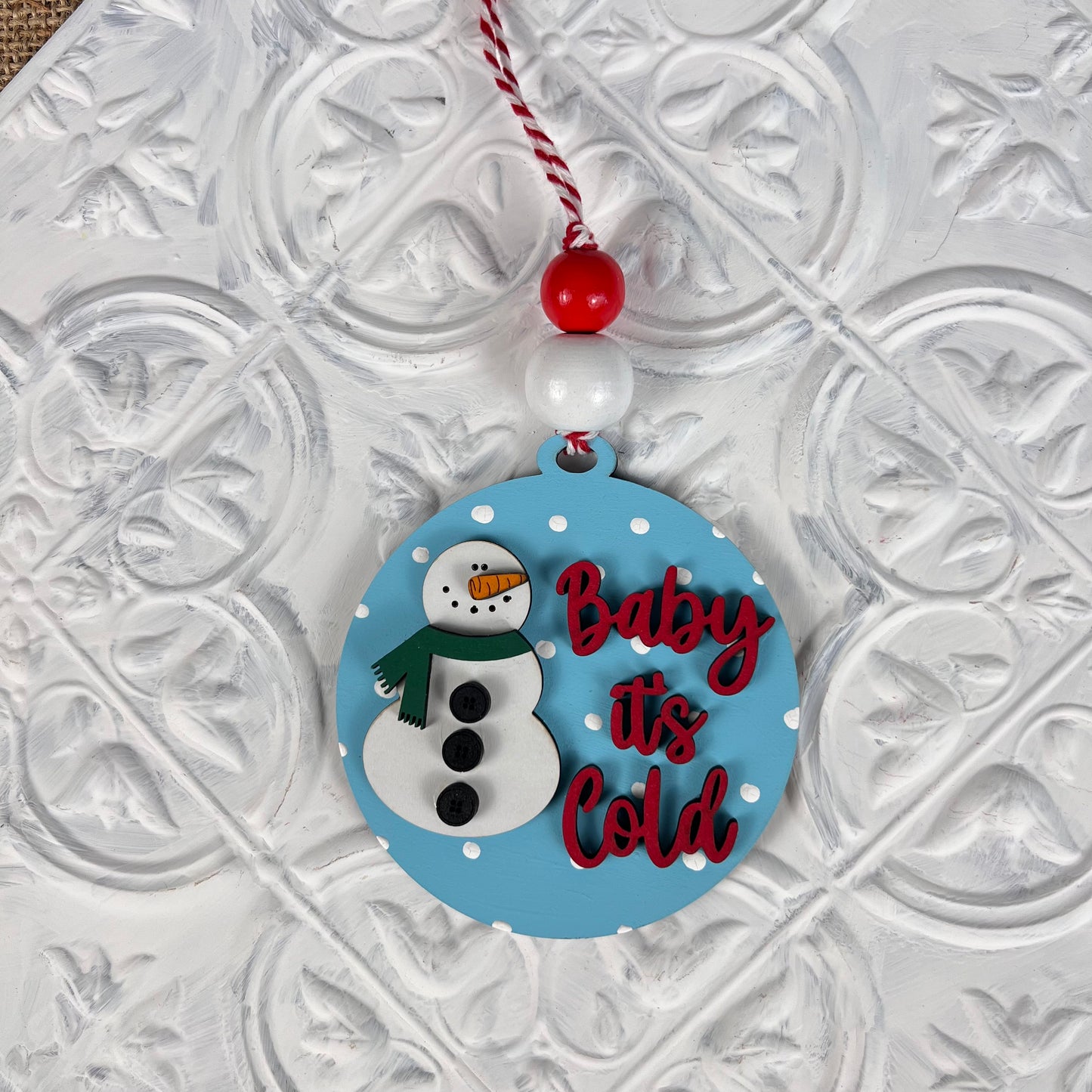 A round Christmas ornament from Janet's Craft Corner's DIY Decoration Kits, featuring a blue design with a snowman and red text stating "Baby it's Cold," hangs from a festive red and white string. This charming decoration, perfect for holiday home décor, is adorned with white polka dots against a textured white background.