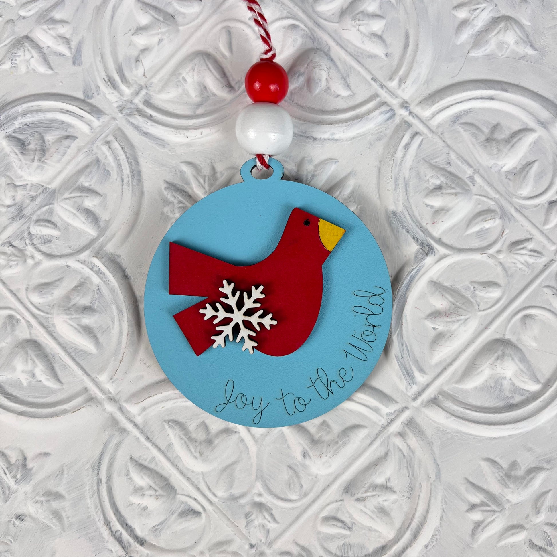 A round Christmas ornament from Janet's Craft Corner's DIY Decoration Kits features a charming light blue design with a red bird and white snowflake decoration. The cursive text "Joy to the World" enhances its appeal. It hangs gracefully from a red and white string, set against an embossed white background—perfect for adding to your DIY kit collection.