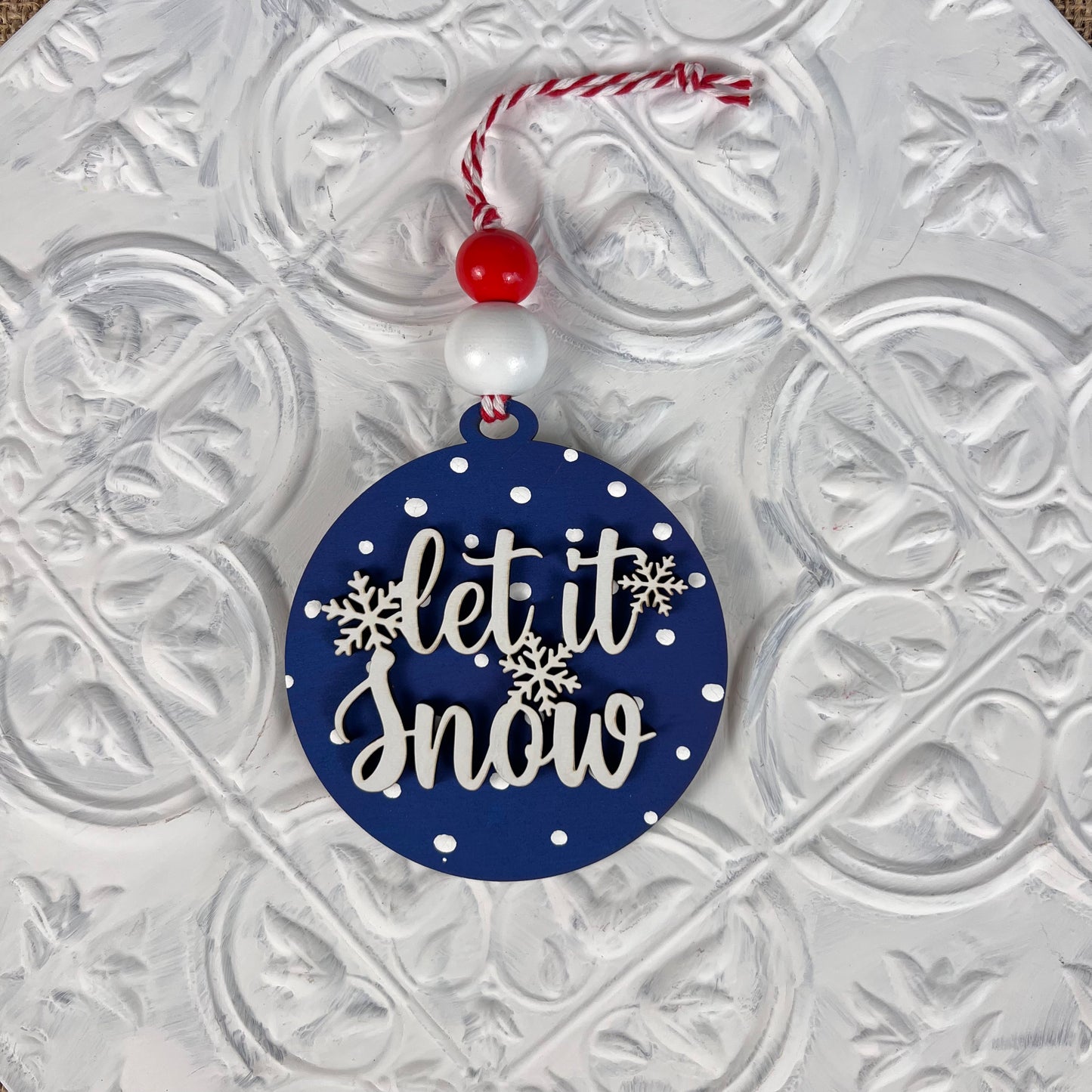 Elevate your festive decorations with Janet's Craft Corner's Round Christmas Ornament DIY Decoration Kit. This lovely round blue ornament, adorned with white polka dots and snowflakes, displays the cheerful message "let it snow." It elegantly hangs from a red and white string embellished with matching beads, making it a charming addition to your decorative white surface.