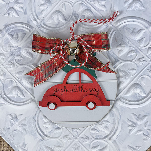 The Jingle All the Way Volkswagen Ornament by Janet's Craft Corner features a delightful red car topped with a festive Christmas tree, all elegantly tied with a red and gold plaid bow and accented with a silver bell, set against a textured white background.