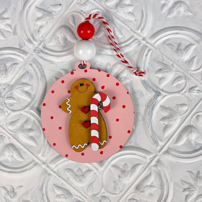 Add a charming touch to your home décor with the Round Christmas Ornaments from Janet's Craft Corner. This captivating ornament features a round pink design adorned with red polka dots and showcases a cheerful gingerbread figure holding a candy cane. Accented with red and white beads and finished with a striped hanger, it beautifully complements any textured white background.