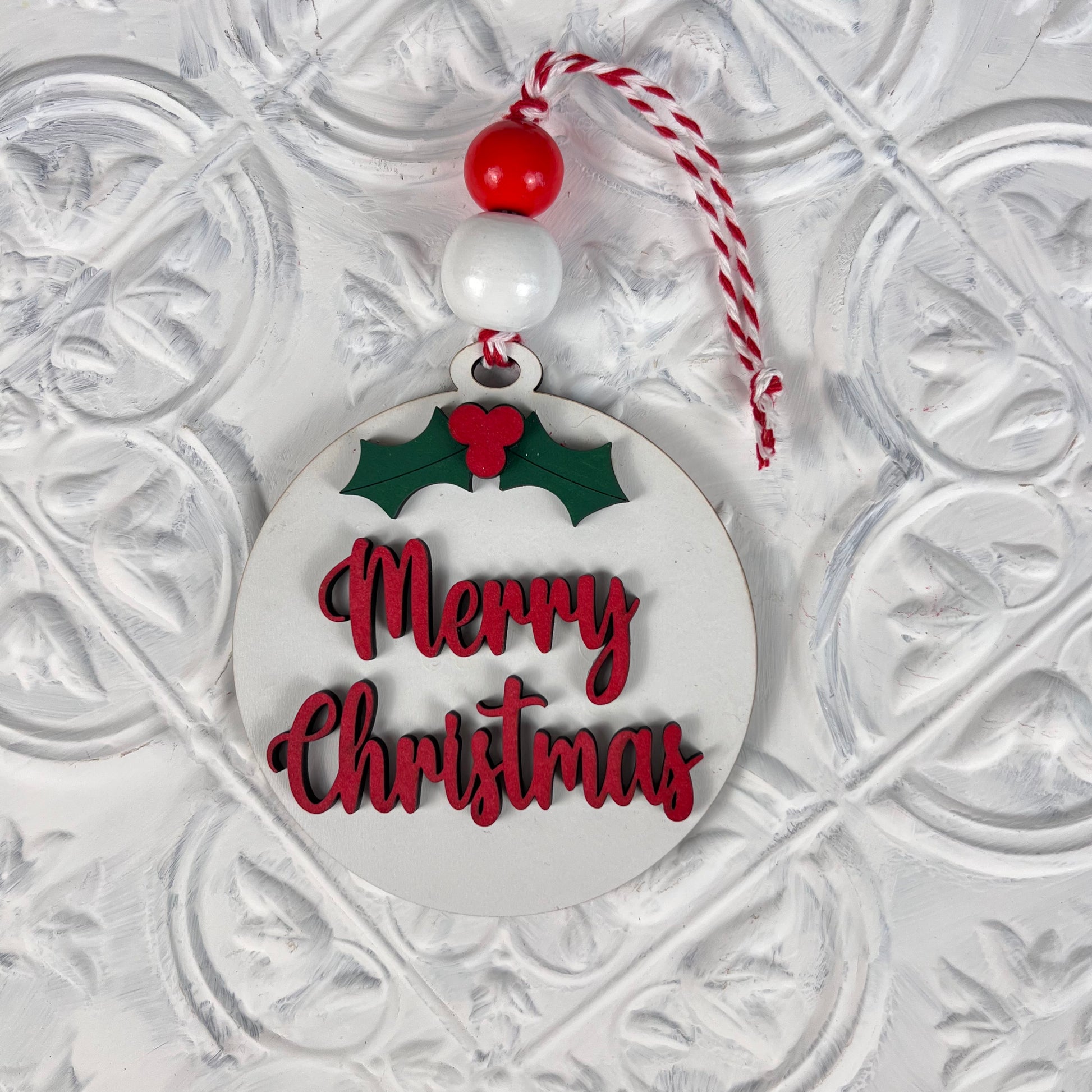 Introducing the Round Christmas Ornaments from Janet's Craft Corner, featuring a charming design with "Merry Christmas" in vibrant red letters. These ornaments include a lovely holly decoration, accented by a decorative red and white bead, and are suspended from a matching red and white string against a festive white background—ideal for enhancing your DIY Decoration Kits this holiday season.