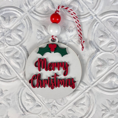 Introducing the Round Christmas Ornaments from Janet's Craft Corner, featuring a charming design with "Merry Christmas" in vibrant red letters. These ornaments include a lovely holly decoration, accented by a decorative red and white bead, and are suspended from a matching red and white string against a festive white background—ideal for enhancing your DIY Decoration Kits this holiday season.