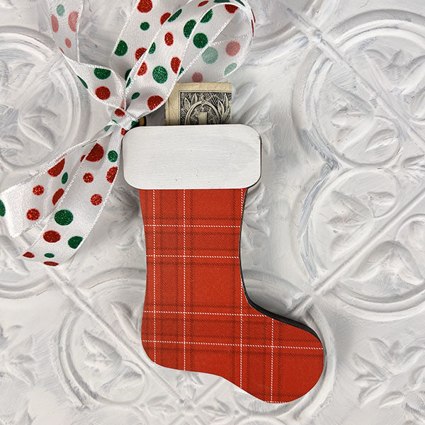 The Stocking Money Holder Ornament - Painted from Janet's Craft Corner is a delightful red plaid Christmas stocking-shaped ornament, perfect for a holiday cash gift. It hangs from a white ribbon adorned with red and green dots, with a dollar bill peeking from the top against a textured white background.